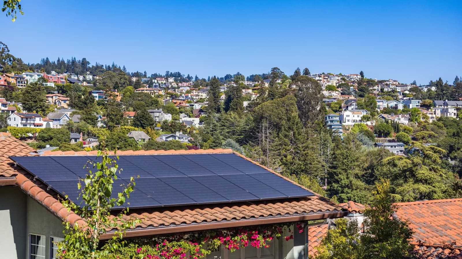 A tiled suburban roof carrying a solar array, with houses on a wooded hillside behind it.
