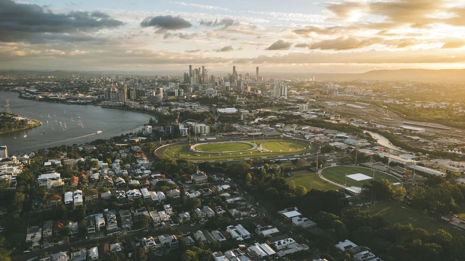 Aerial photograph of Brisbane at sunset, looking over suburban rooftops and the Brisbane River towards the city skyline.