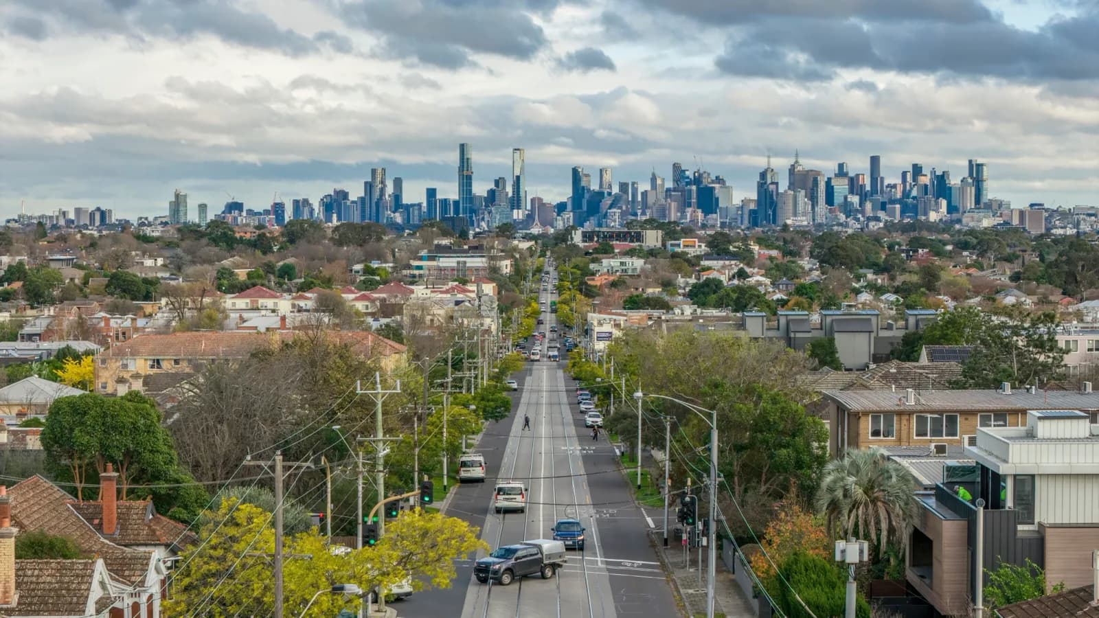 A tree-lined Melbourne suburban street with tram tracks, looking towards the city skyline.