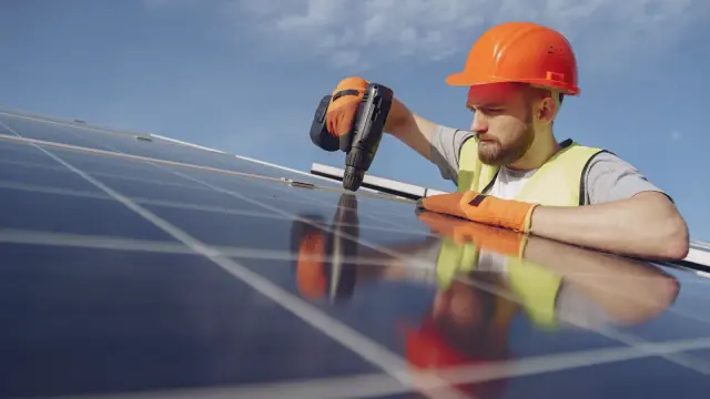 An installer in a hard hat and high-visibility vest fixing a solar panel into place against a blue sky.