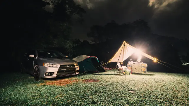 A car beside camping tents and a lit shelter on a grassy campsite at night.
