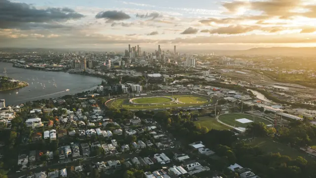 Aerial photograph of Brisbane at sunset, looking over suburban rooftops and the Brisbane River towards the city skyline.
